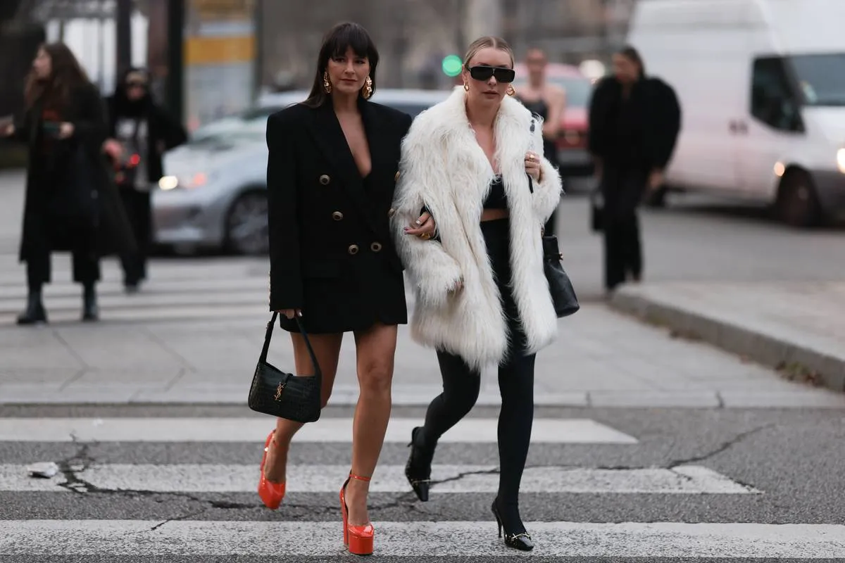 PARIS, FRANCE - JANUARY 24: A Fashion Week guest wearing a black oversized long blazer, gold statement earrings and a YSL black leather bag and Justyna Czerniak wearing a black sunglasses, gold earrings, a black cropped Christian Dior bra, white oversized fake fur coat, a black shiny leather quilted pattern shoulder bag from Saint Laurent, black nylon pants, black shiny varnished leather with gold metallic embroidered toe-cap heels shoes is seen wearing before the Alexandre Vauthier show on January 24, 2023 in Paris, France. 